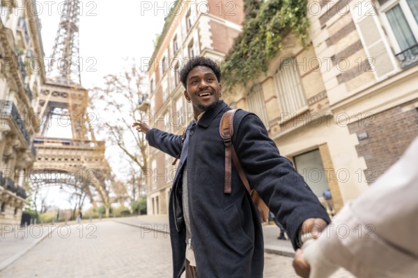 Happy young man of african descent leads his partner by the hand, pointing toward the eiffel tower while smiling during a romantic city break in paris, france