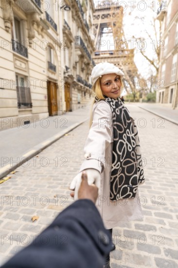 Woman holding her partner's hand, smiling and looking back while walking on a cobblestone paris street with haussmann buildings and the eiffel tower in the background, romantic travel vibe
