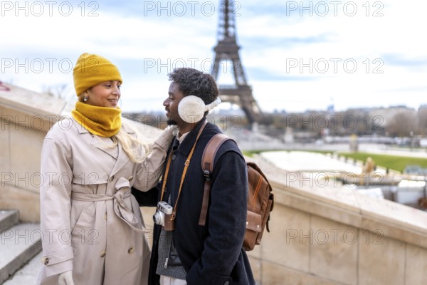 Diverse couple standing together with the eiffel tower in the background, enjoying their romantic trip to the famous european city during winter and wearing warm clothing