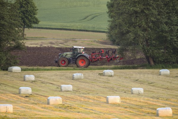 A tractor plows a field in a rural landscape with round bales of hay bales in the foreground, Osnabrücker Land, Lower Saxony, Germany