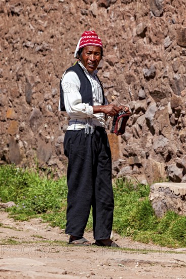 Man wearing a red cap and traditional clothing walks along a stone wall, The knitting men on Taquile Island in Lake Titicaca in Peru