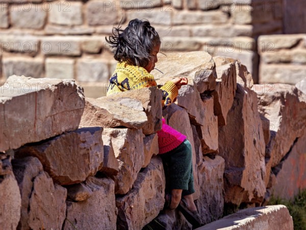 Little girl in colorful dress sitting on a stone wall and smiling, children from Taquile Island in Lake Titicaca in Peru