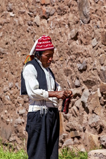 Close-up of a man wearing a red cap and traditional clothes on a stone wall, The knitting men on the island of Taquile in Lake Titicaca in Peru