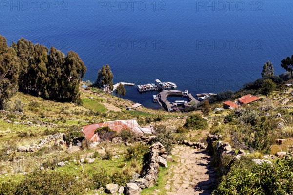 Trail leads to boats on the shores of a deep blue lake surrounded by vegetation, countryside and farmhouses on the island of Taquile in Lake Titicaca in Peru
