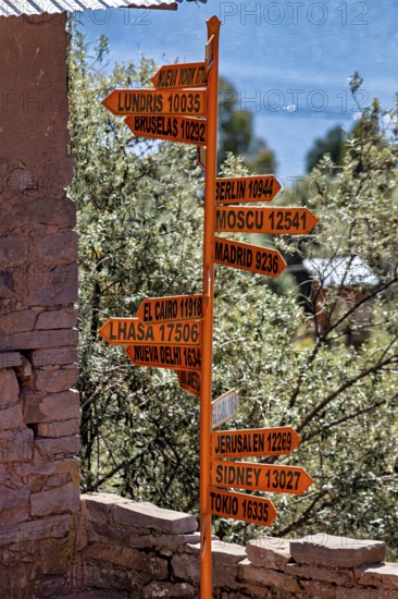 An orange signpost with several international cities and distances in front of a stone wall in a natural setting, signposts of the capitals on the island of Taquile in Lake Titicaca in Peru