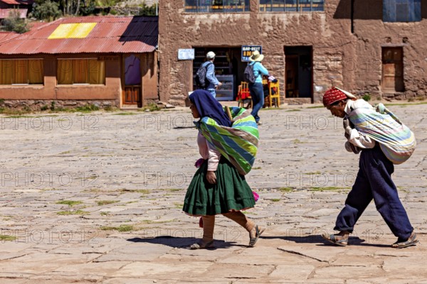 Two woman in traditional clothing carry bundles on their backs through a village in Bolivia in daylight, farmers walk across a square on the island of Taquile in Lake Titicaca in Peru