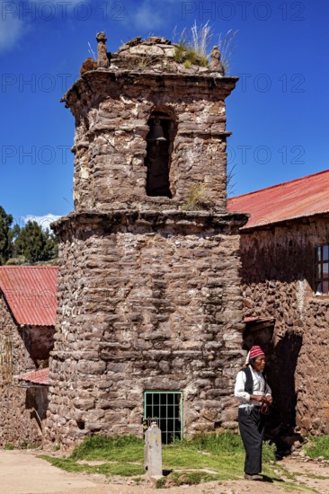 Stony church tower with red roof and a man wearing traditional clothes next to it, The knitting men on the island of Taquile in Lake Titicaca in Peru