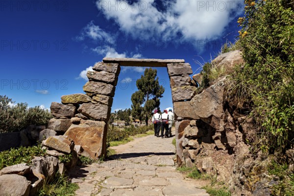 People wearing traditional clothes walk under a rectangular stone arch on a rural path, stone gates on Taquile Island in Lake Titicaca in Peru