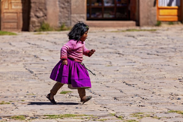Little girl in purple skirt and pink sweater walks across a paved square, children from Taquile Island in Lake Titicaca in Peru