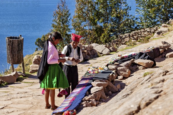 People wearing traditional clothing sell goods on a rock path, overlooking a large lake, the woman on Taquile Island in Lake Titicaca in Peru