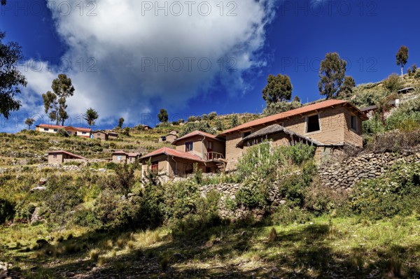 Houses cluster on a hillside under a dramatically cloudy sky framed by trees and vegetation, The countryside and farmhouses on the island of Taquile in Lake Titicaca in Peru