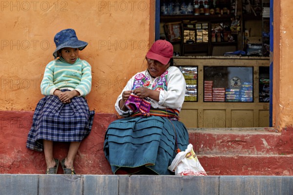 Two woman in traditional clothes sit on a staircase next to a small shop, The woman on Taquile Island in Lake Titicaca in Peru