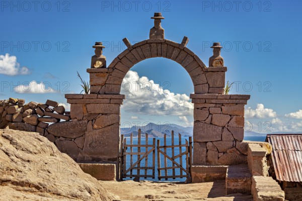 A traditional stone arch with wooden slats stands in front of a distant mountain landscape under clear blue skies, stone gates on the island of Taquile in Lake Titicaca in Peru