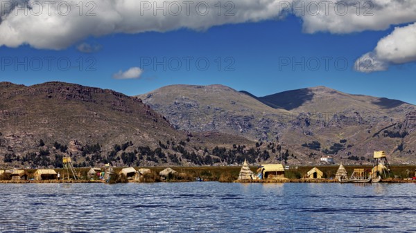 Reed huts on the shores of a tranquil lake in front of a vast mountain landscape under a blue sky, The floating reed islands of the Uros in Lake Titicaca in Peru