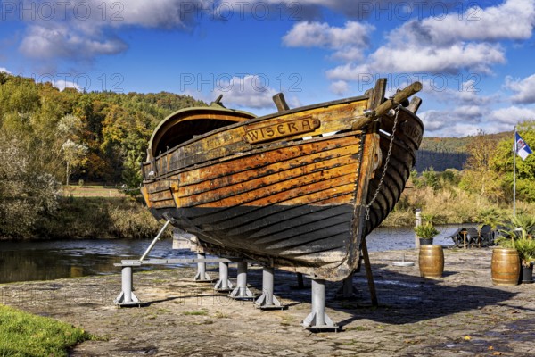 Historic wooden boat on riverbank, surrounded by autumn trees under a clear sky, the port of Wanfried an der Werra with the historic Werra ship