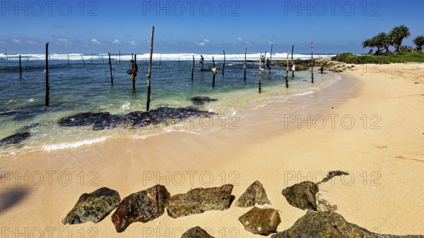 Quiet beach with clear waves and wooden poles under blue skies, Koggala beach with stilt fishermen in Sri Lanka