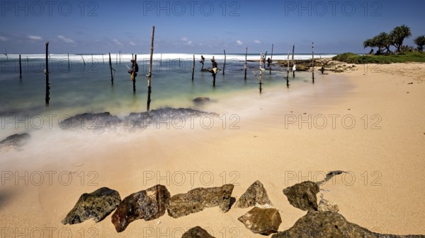 Clear beach with visible rocks and wooden posts in the water, Koggala beach with stilt fishermen in Sri Lanka