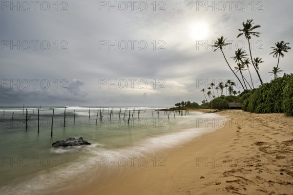 Tropical beach with soft waves, palm trees and cloudy sky, Koggala beach with stilt fishermen in Sri Lanka