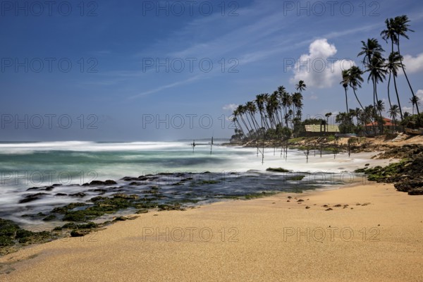 A sun-drenched beach with palm trees and waves hitting the rocks, Koggala beach with stilt fishermen in Sri Lanka