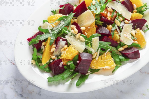 A salad made with chunks of beet, orange slices, and arugula. It is topped with cheese shavings and pine nuts, placed on a white plate with a striped napkin beside it