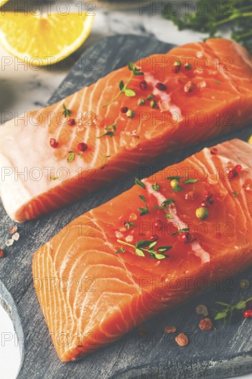 Two pieces of salmon fillet sit on a wooden cutting board. They are topped with herbs and pink peppercorns. Slices of lemon and lime surround the fish, creating a vibrant setting for cooking