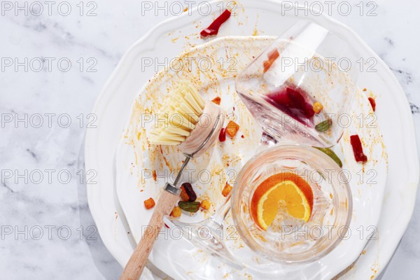 Dishes are stacked on a kitchen counter with food remnants after a meal. A cleaning brush and sponge are ready for washing the plates and glasses
