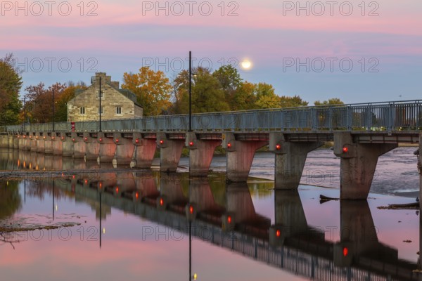 Illuminated Moulin Neuf water flow control dam and walkway over Des Mille-Iles river plus New Mill on Ile des Moulins in autumn at dusk, Old Terrebonne, Quebec, Canada