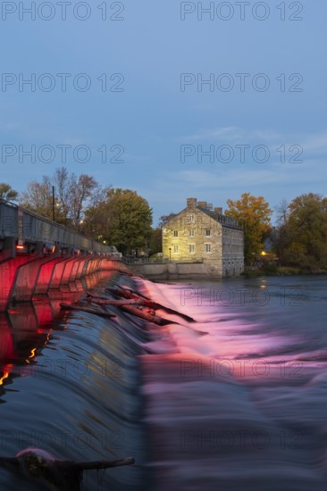 Illuminated Moulin Neuf water flow control dam and walkway over Des Mille-Iles river plus New Mill on Ile des Moulins in autumn at dusk, Old Terrebonne, Quebec, Canada