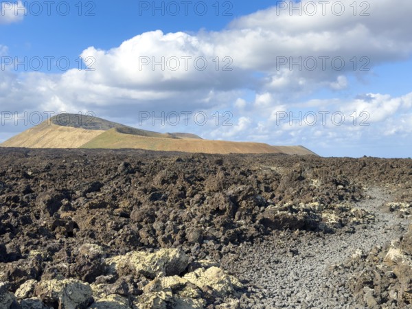 Hiking trail through lava rock to Caldera Blanca volcano against blue sky with white clouds, Mancha Blanca, Lanzarote, Canary Islands, Spain