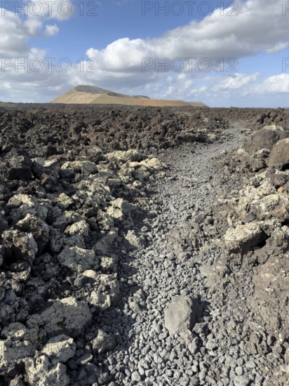 Hiking trail through lava rock to Caldera Blanca volcano against blue sky with white clouds, Mancha Blanca, Lanzarote, Canary Islands, Spain