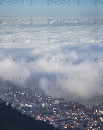 Aerial view of the city of Heidelberg on the Neckar, sea of fog from low-hanging clouds and fog fields, sunlit above the roofs of the city and the Neckar river, floating and waving with large and smoking chimney vents sticking out of the fog in the background, Heidelberg, Baden-Württemberg, Germany