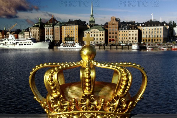 A golden crown in the foreground with a view of the skyline and boats of Stockholm, Stockholm, Gamla Stan, Sweden