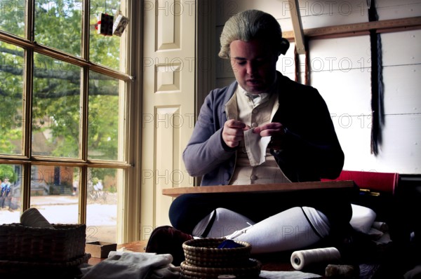 A tailor in period clothing works concentrated in a well-lit room, Williamsburg, Virginia, USA