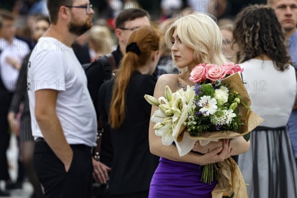 Blonde woman holding a bouquet of flowers during a high school graduation ceremony in a busy square, Vilnius, Vilnius County, Lithuania