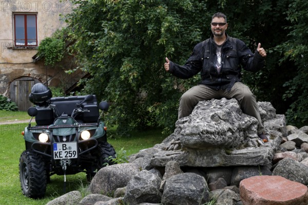 Person sitting on a crocodile sculpture next to an ATV in a green setting, Dundaga, Latvia