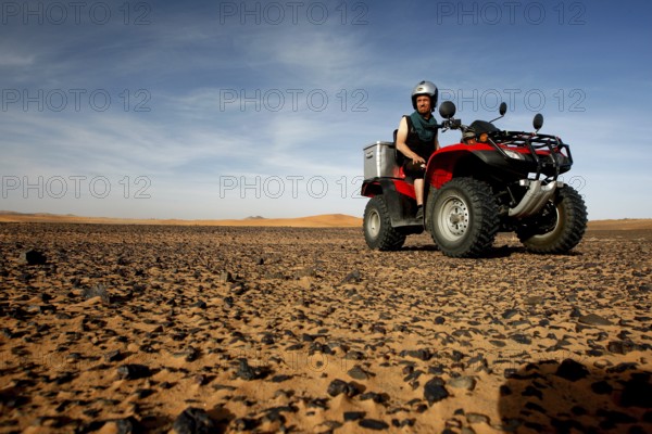 A red quad bike rides across a rocky desert landscape under a blue sky, Erg Chebbi, Hammada, Morocco
