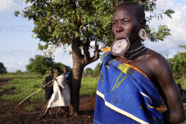 Mursi woman with lip plate in front of a tree in Mago National Park. A village can be seen in the background, Mago National Park, Ethiopia