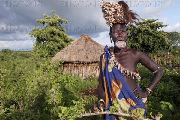 Mursi woman with lip plate in front of a hut in Mago National Park. The landscape is green and the sky is cloudy, Mago National Park, Ethiopia