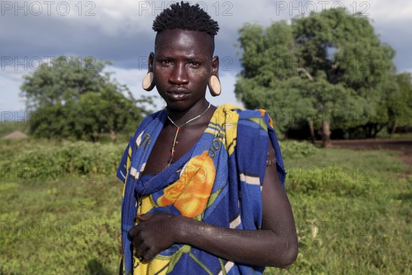 Mursi man wearing traditional clothing and earrings in front of lush landscape in Mago National Park, Mago National Park, Ethiopia