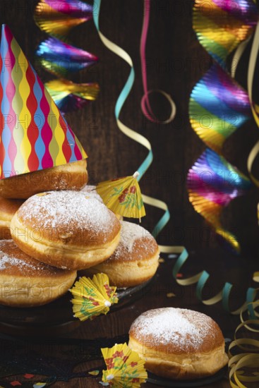 A pile of donuts with powdered sugar, colorful garlands and party hat against a dark wooden background, carnival