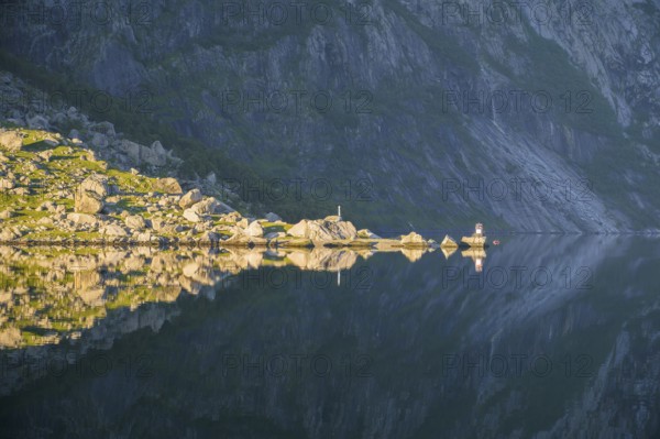 Sunny rocky landscape reflected on the calm water surface of Lysefjord, calm water reflects rocks and stones under a blue sky, Lysefjord, Rogaland, Norway
