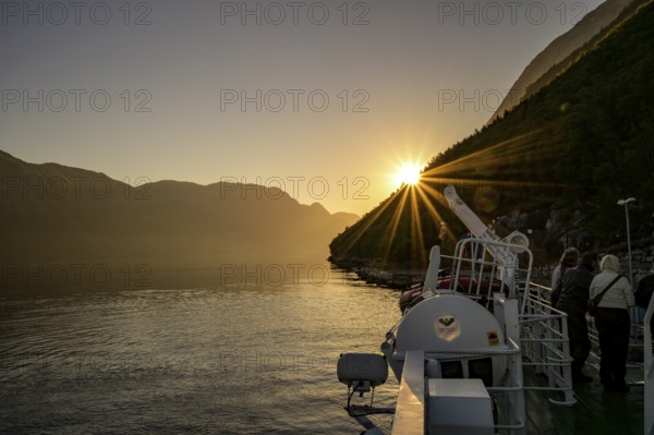 A boat sails through the Lysefjord at sunset with sun star with calm water and surrounded by mountains, view from the Columbus ferry, Lysefjord, Rogaland, Norway