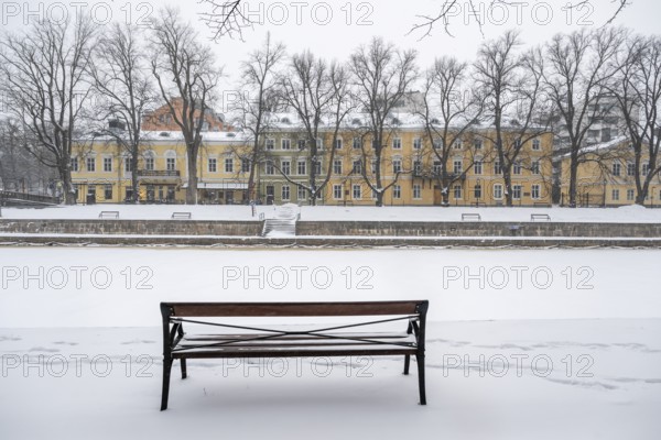 Bench and historic buildings on the frozen Aurajoki river, wintery Turku or Swedish Åbo, Varsinais-Suomi, Finland
