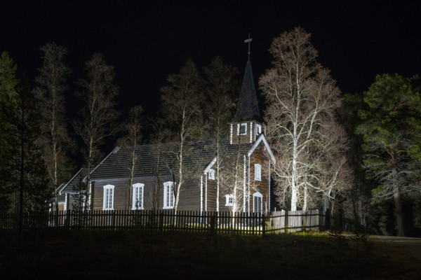 An illuminated church stands at night surrounded by trees and a wooden fence