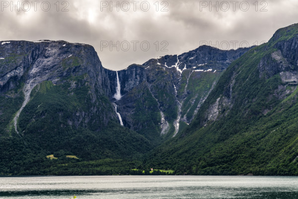 A waterfall flows between steep mountains into a quiet fjord under a cloudy sky