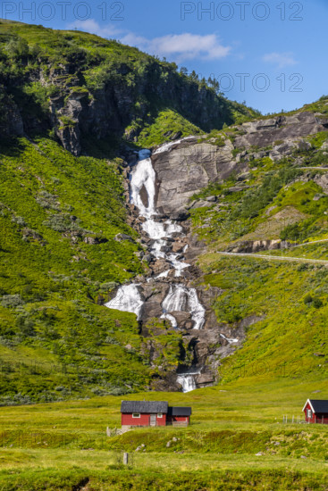 A cascading waterfall flows over green hills next to a red hut in the sunshine