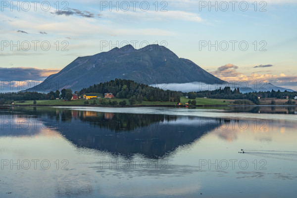 A mountain is reflected in a peaceful lake, surrounded by meadows and fields under an evening sky