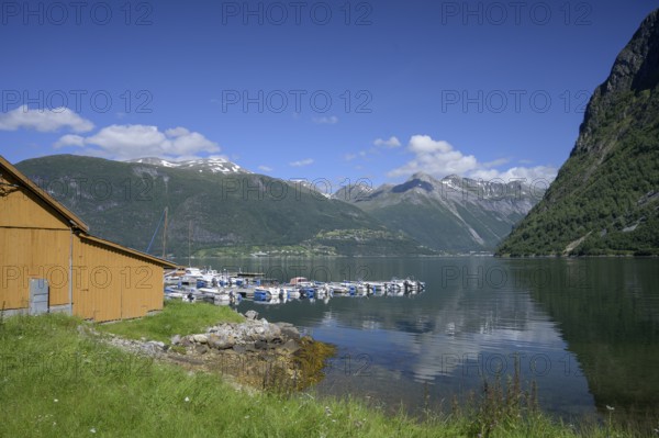 Norddal, Møre og Romsdal province, Norway, wooden cabin on the shore of a fjord with boats and mountains in the background under blue sky
