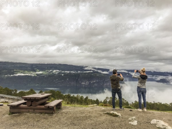 Utvik, Vestland, Norway, Two people taking pictures of a misty mountain backdrop with a lake in the valley on their smartphones