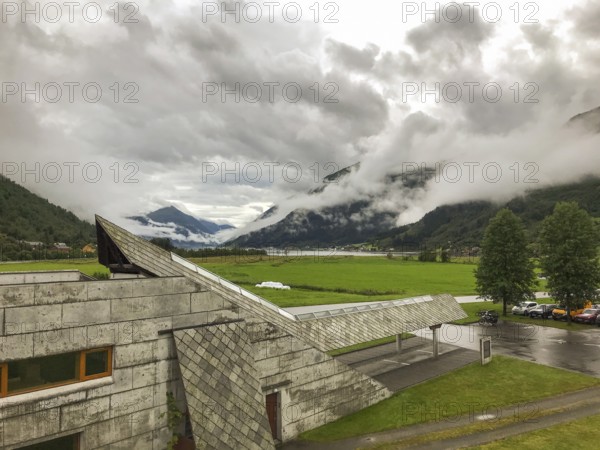Fjærland, Vestland, Norway, concrete building of the Norwegian Glacier Museum Norsk Breecenter in a green valley on the Jostedalsbreen glacier, surrounded by cloudy mountains after rain, modern building between fog covered mountains with green landscape in the valley